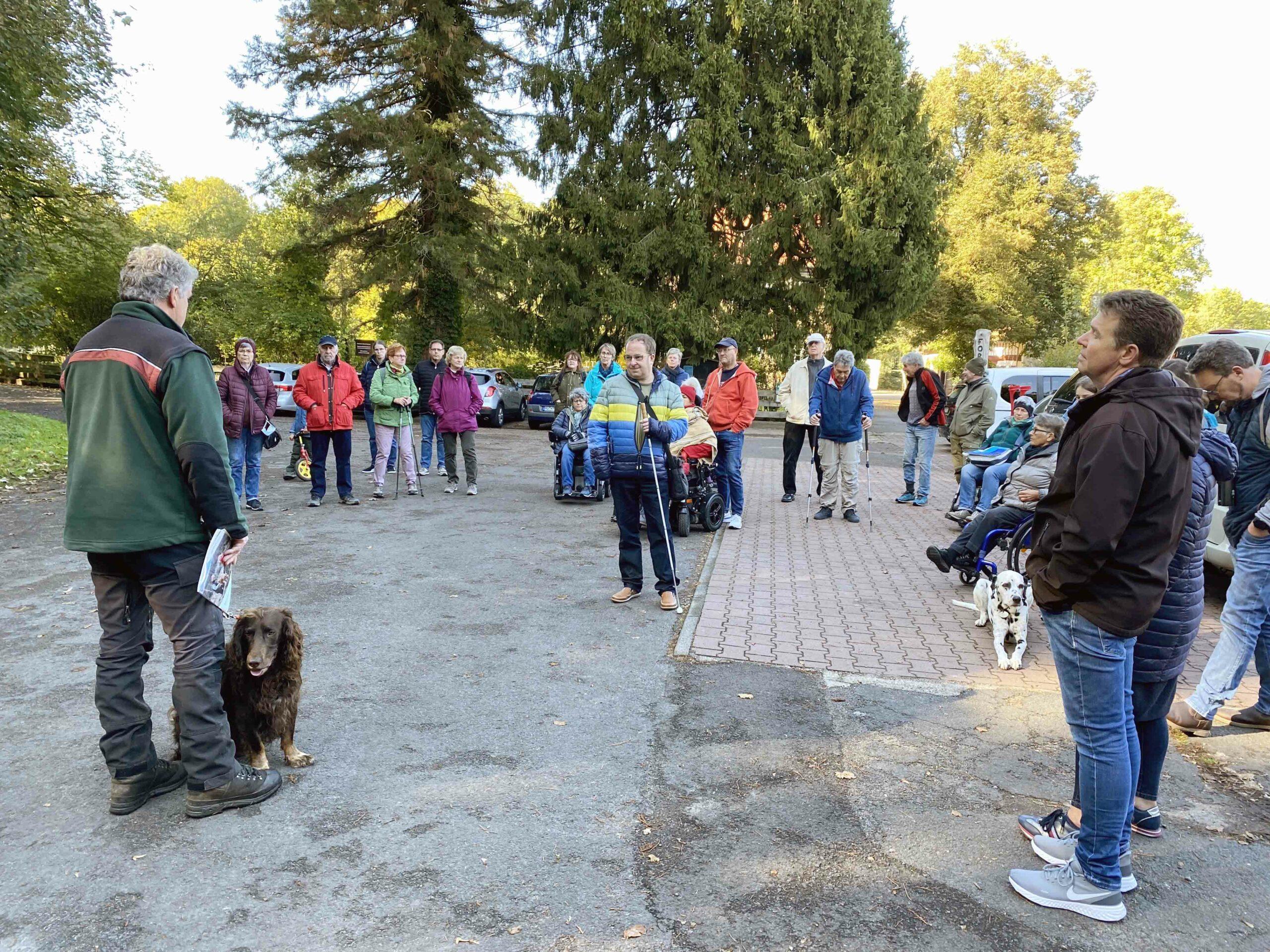 Blick auf eine Gruppe von ca. 25 bis 30 Menschen von ganz jung bis Älter mit und ohne Hilfsmitteln. Links im Bild ist der Förster zu sehen, mit seinem Hund an der Leine.