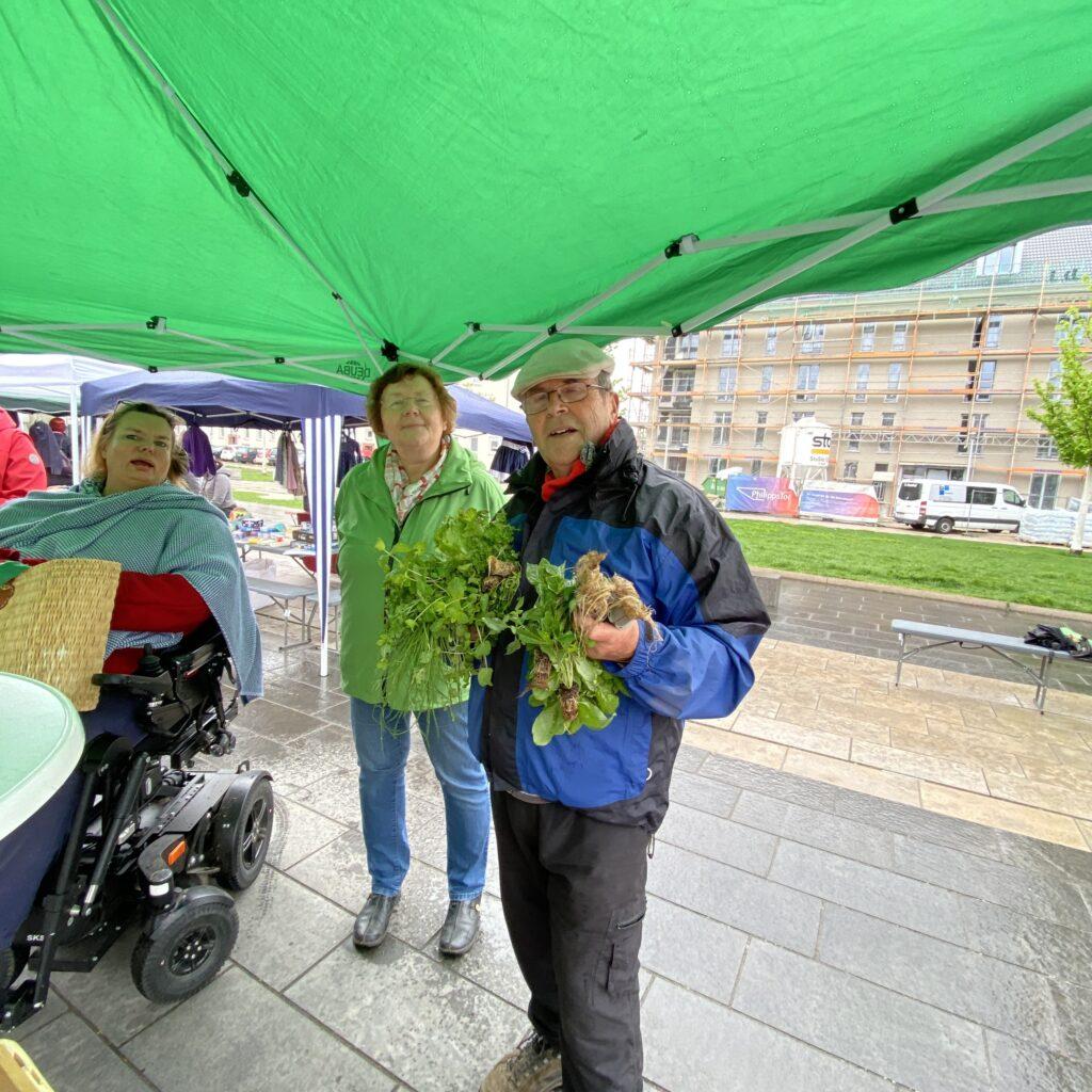 Eine Person im Rollstuhl und ein Mann und eine Frau stehen unter einem grünen Zeltdach auf dem Platz.