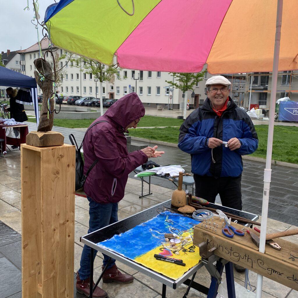 Ein Mann mit Kunststücken aus Holz steht unter einem bunt gestreiften Schirm.