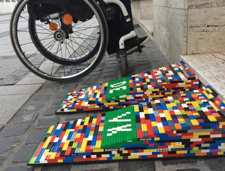 Man sieht die hohe zweispurige LEGO-Rampe mit DEVK in weißen LEGO-Steinen auf grünem Grund. Dahinter steht ein Rollstuhl.