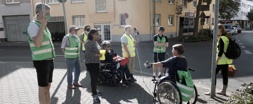 Eine Gruppe von Menschen in Warnwesten, die vor einem Zebrastreifen stehen. Dort erklärt eine Frau im Rollstuhl Regeln.