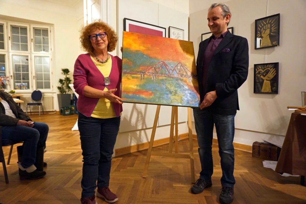Rina Nentwig übergibt ihr Kunstwerk an den Mann, der ihr Kunstwerk ersteigert hat. Sie steht links er rechts. Sie halten beide das Bild fest.