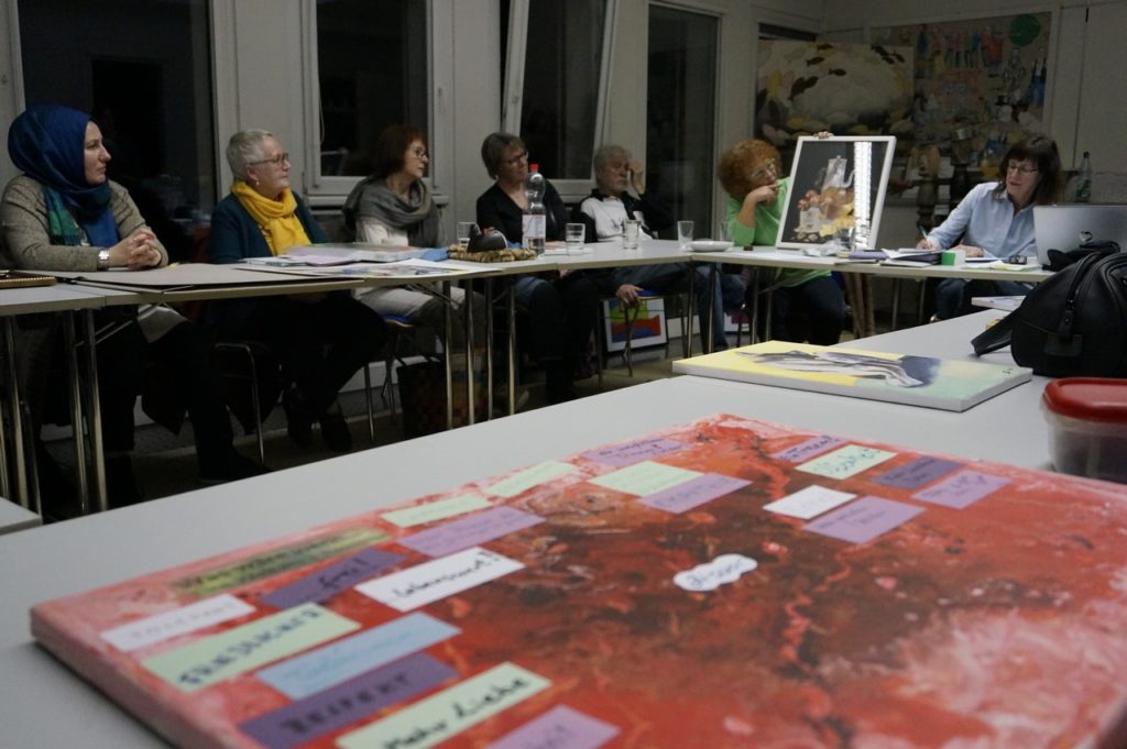 Mehr als 30 Menschen haben sich an unserer Aktion beteiligt. Im Vordergrund liegt ein Kunstwerk auf dem Tisch. Im Hintergrund erkennt man einen Teil der Gruppe an Tischen sitzend.