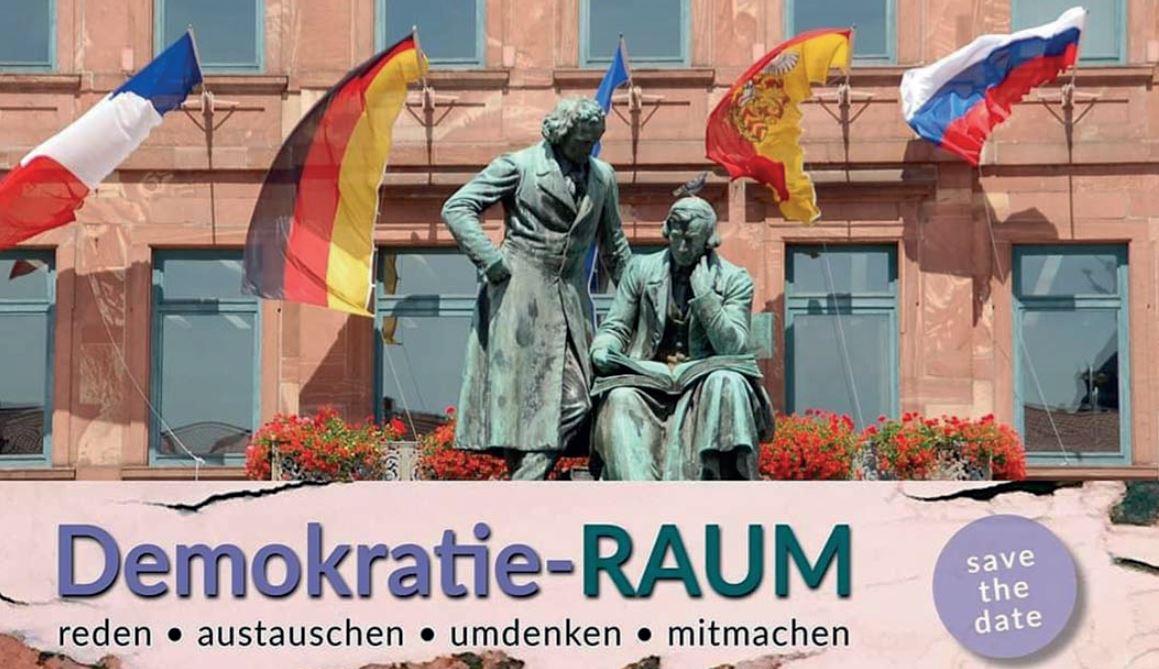 Das Bild zeigt im Hintergrund das Brüder Grimm Denkmal vor dem Rathaus am Marktplatz. Davor in großen Lettern Demokratie-RAUM mit der Erklärung für die einzelnen Buchstaben: reden, austauschen, umdenken, mitmachen.