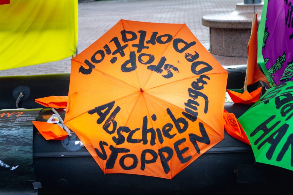 Aufgespannter Regenschirm am Schlauchboot von AK Asyl. Er trägt die Schrift Abschiebungen stoppen und Stop Deportation.