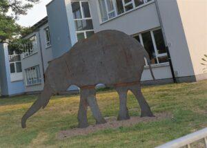 Ein grauer, circa 2 Meter hoher, 2-dimensionaler Elefant aus Stahl steht für einem Schulgebäude