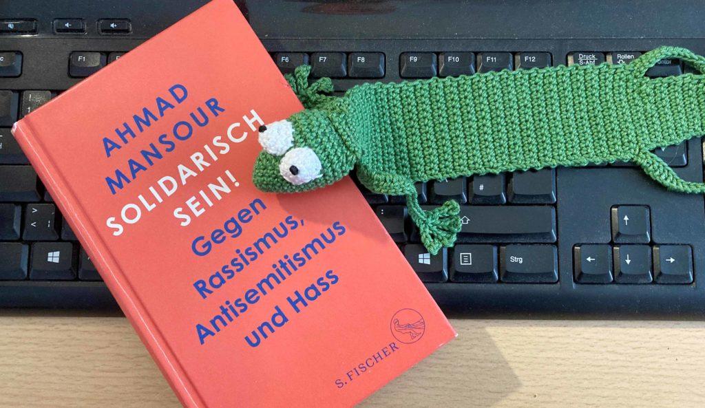 Das Buch von Ahmad Mansour "Solidarisch sein!" mit rotem Bucheinband liegt auf einer schwarzen Tastatur. Daneben liegt ein Buchlesezeichen gehäkelt in Form eines grünen Frosches.