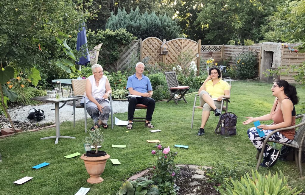 Vier Menschen sitzen im Kreis mit großen Abstand in einem blühenden Garten. Auf dem Gras liegen bunte Zettel mit Stichworten.