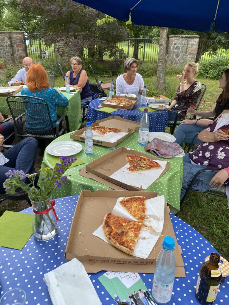 Menschen sitzen um Tische herum in einem Garten und verspeisen Pizzastücke.
