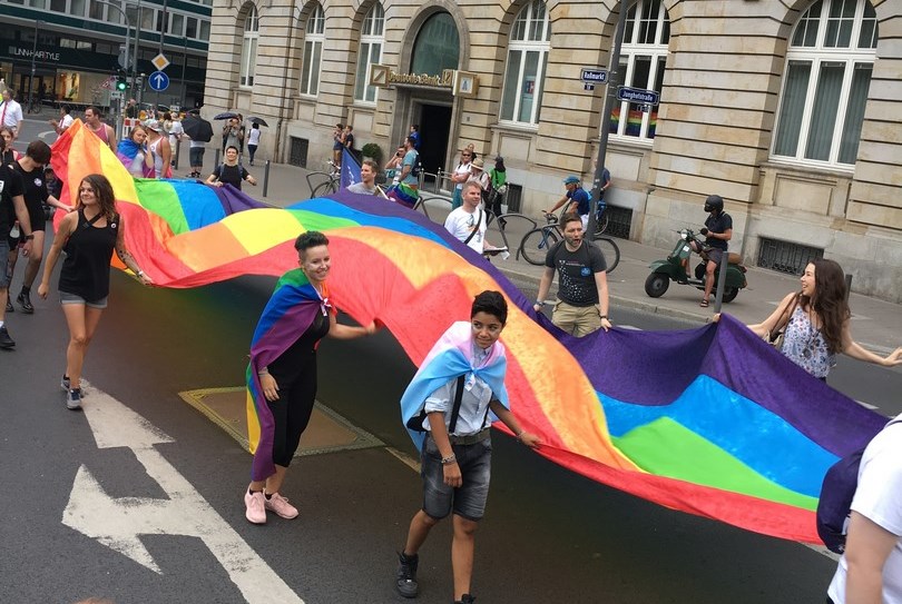 Zu Beginn wird eine große Regenbogen-Flagge getragen. Etwa 20 Menschen tragen diese parallel zum Boden und machen ab und an eine Laola Welle.