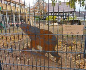 In der Seitenansicht ist ein laufender Bär zu sehen, der auf dem Schulhof der Montessorie-Schule steht. Vor dem Bären ist ein Gitter zu sehen, weil die Schule sich gerade in Renovierung befindet.
