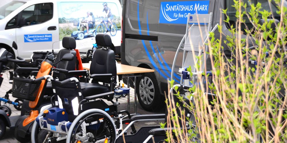 Rollstühle, Rollatoren und andere Hilfsmittel sind aufgereiht vor einem Sprinter des Sanitätshauses Marx