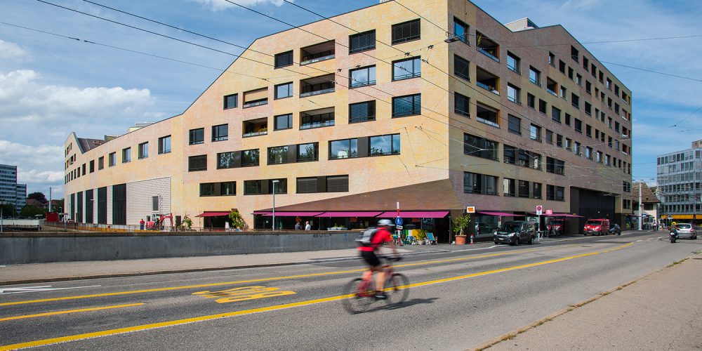 Ein mehrstöckiger Gebäudekomplex, davor eine Straße mit einem Radfahrer und Autos.