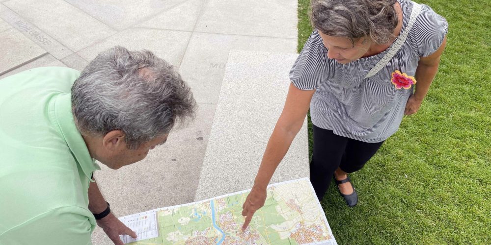 Susanne zeigt Peter wo sich eine öffentliche Toilette befindet. Sie deuten mit den Händen auf eine Karte von Hanau.
