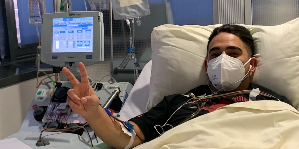 Das Bild zeigt Belal mit einer Maske im Bett in einem Krankenhaus liegend. Stolz hält er mit der rechten Hand das Peace-Zeichen in die Kamera.