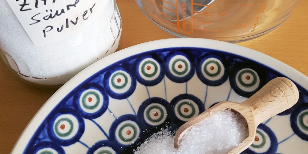 Auf einem Keramikteller mit einem blauen Muster liegt ein kleiner hölzerner Löffel. Darüber Steht rechts ein Flasche mit der Aufschrift Zitronensäure - Pulver. Daneben ist ein Messbecher aus Glas zu sehen.