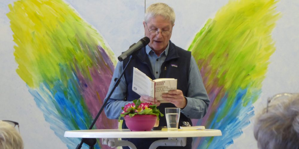 Ein Mann steht vor einem Stehtisch und liest aus einem Buch vor. Im Hintergrund sind bunte Engelsfügel auf einer Wand zu sehen. Der Vorleser steht so, dass die Flügel rechts und links neben seinem Körper sind.