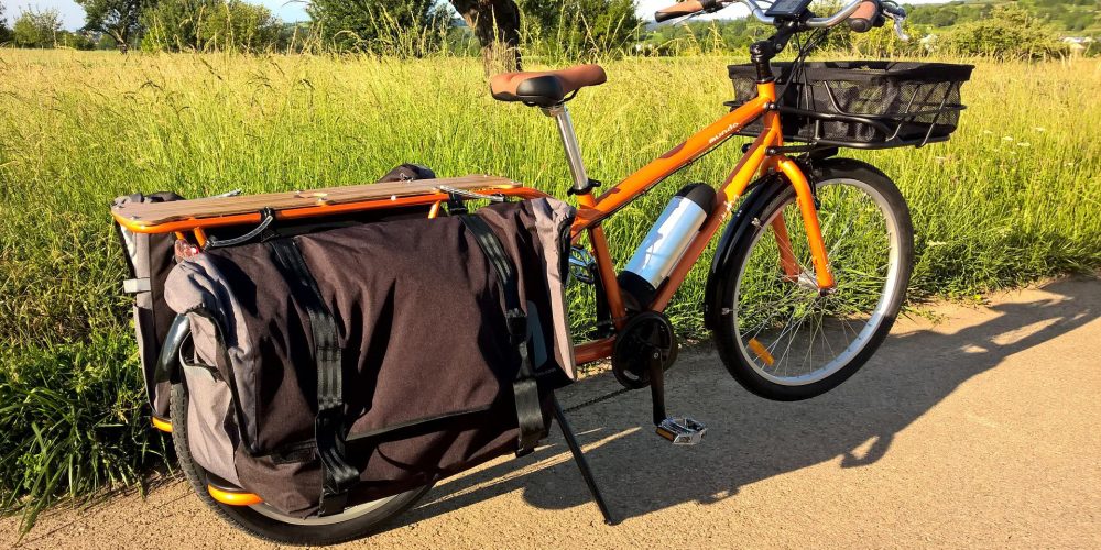 Es ist ein orangenes Lasten-Fahrrad zu sehen das neben einem Feld and einem sonnigen Tag steht.