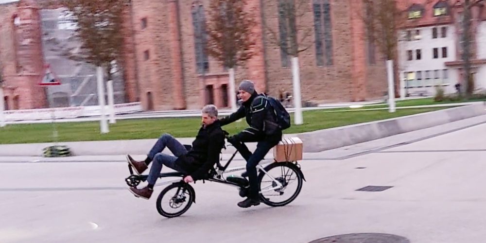 Auf dem Pino-Tandem sitzen 2 Männer und fahren vor der Wallonisch-Niederländischen Kirche