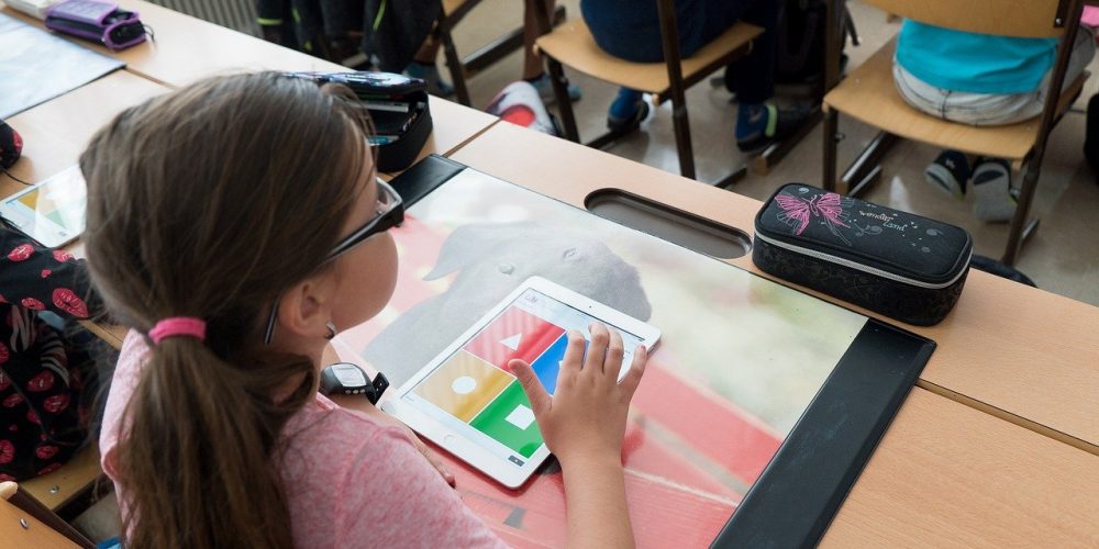 Wir sehen eine Schülerin, die mit einem Tablet am Unterricht teilnimmt. Sie sitzt an einem Holztisch, trägt eine Brille und ein rosafarbenes T-Shirt.