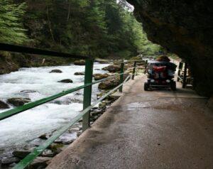 Es ist ein Fluss neben einer Felswand auf der einen und einem Wald auf der anderen Seite zu sehen. Kirsten fährt mit Ihrem Rollstuhl entlang eines befestigten Pfades.