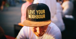 Ein junger Mann mit rötlichem Bart, der eine Baseball-Kappe auf hat, auf der steht "Liebe Deinen Nachbarn". Die Kappe ist im Fokus im Bild.