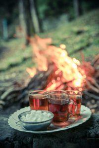 Im Hintergrund brennt ein Feuer auf einer Wiese; davor stehen auf einem kleinem weißen Tellerchen 4 Teegläser und ein Zuckerschälchen.