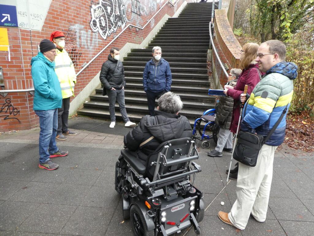 Zu sehen ist die steile Treppe zu den Bahnsteigen des Bahnhofs Steinheim und eine Personengruppe, die angeregt diskutiert.